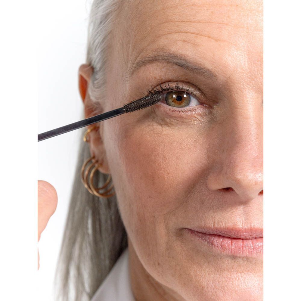 Vrouw met grijs haar, mascara wordt aangebracht. Zwarte borstel. Gouden oorringen.