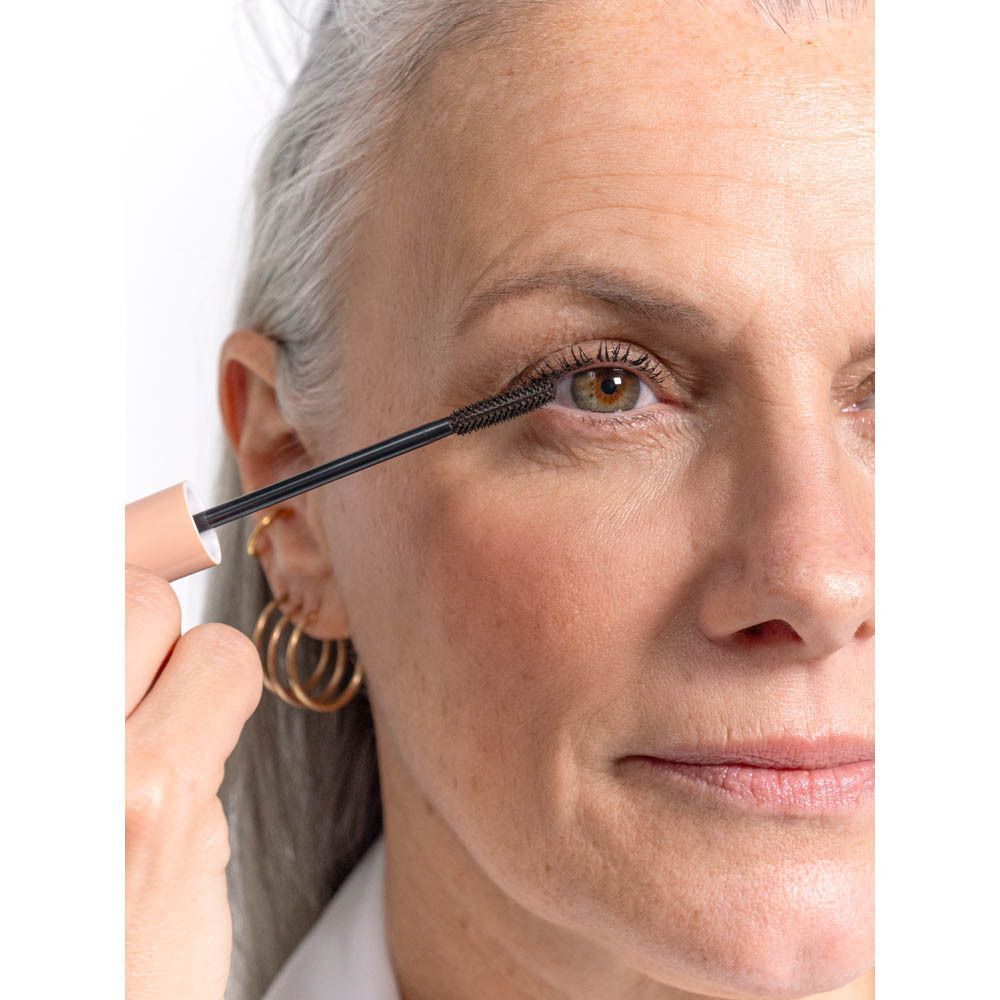 Femme aux cheveux gris, application de mascara. Brosse noire. Créoles dorées.