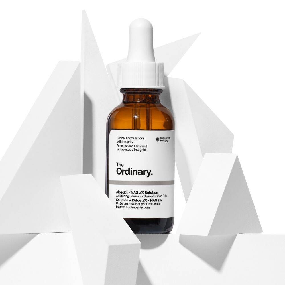 Een flacon van 'The Ordinary Aloe 2% + NAG 2% Solution' met pipet, omringd door witte geometrische vormen.