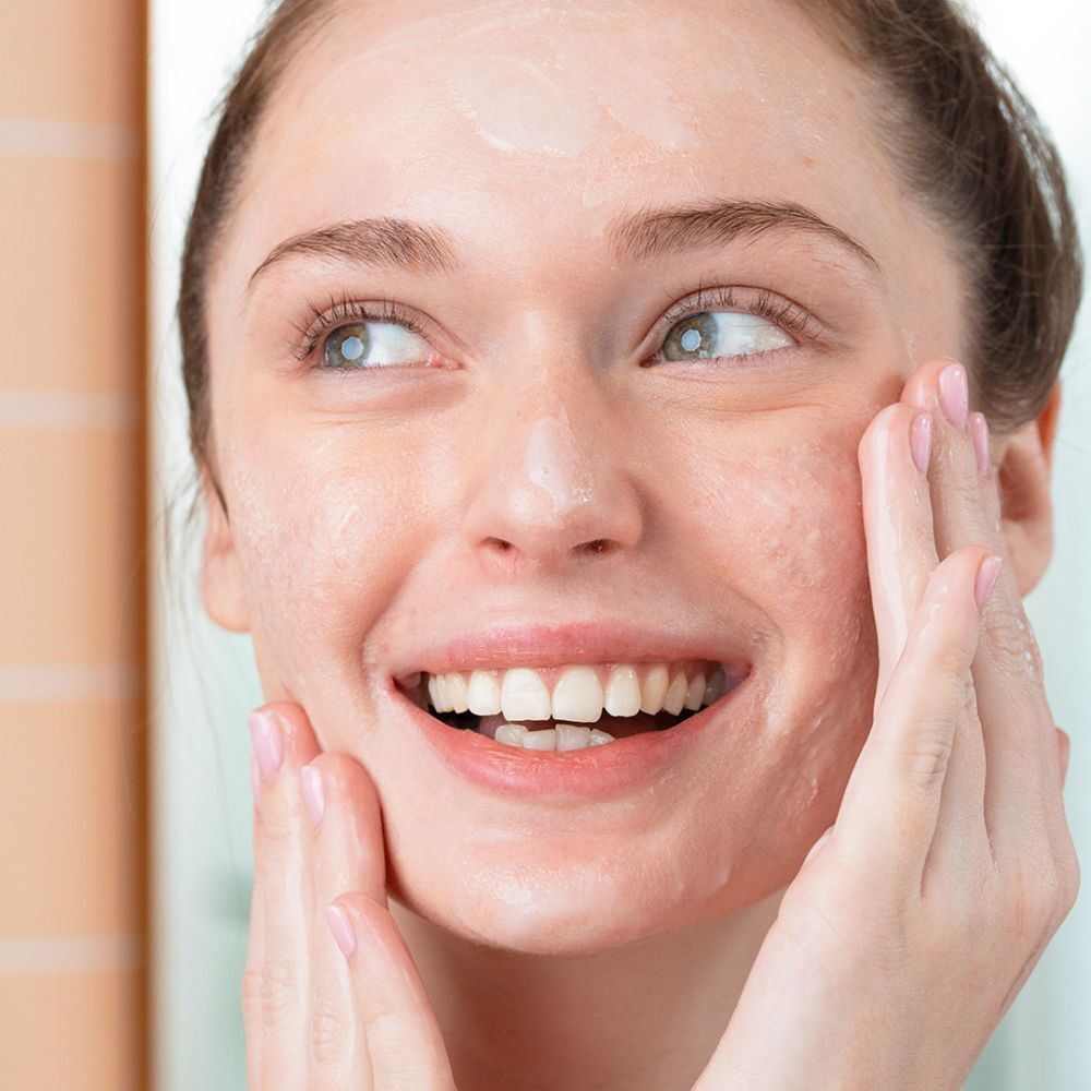 Une femme applique de la crème sur son visage. Elle sourit et regarde sur le côté.