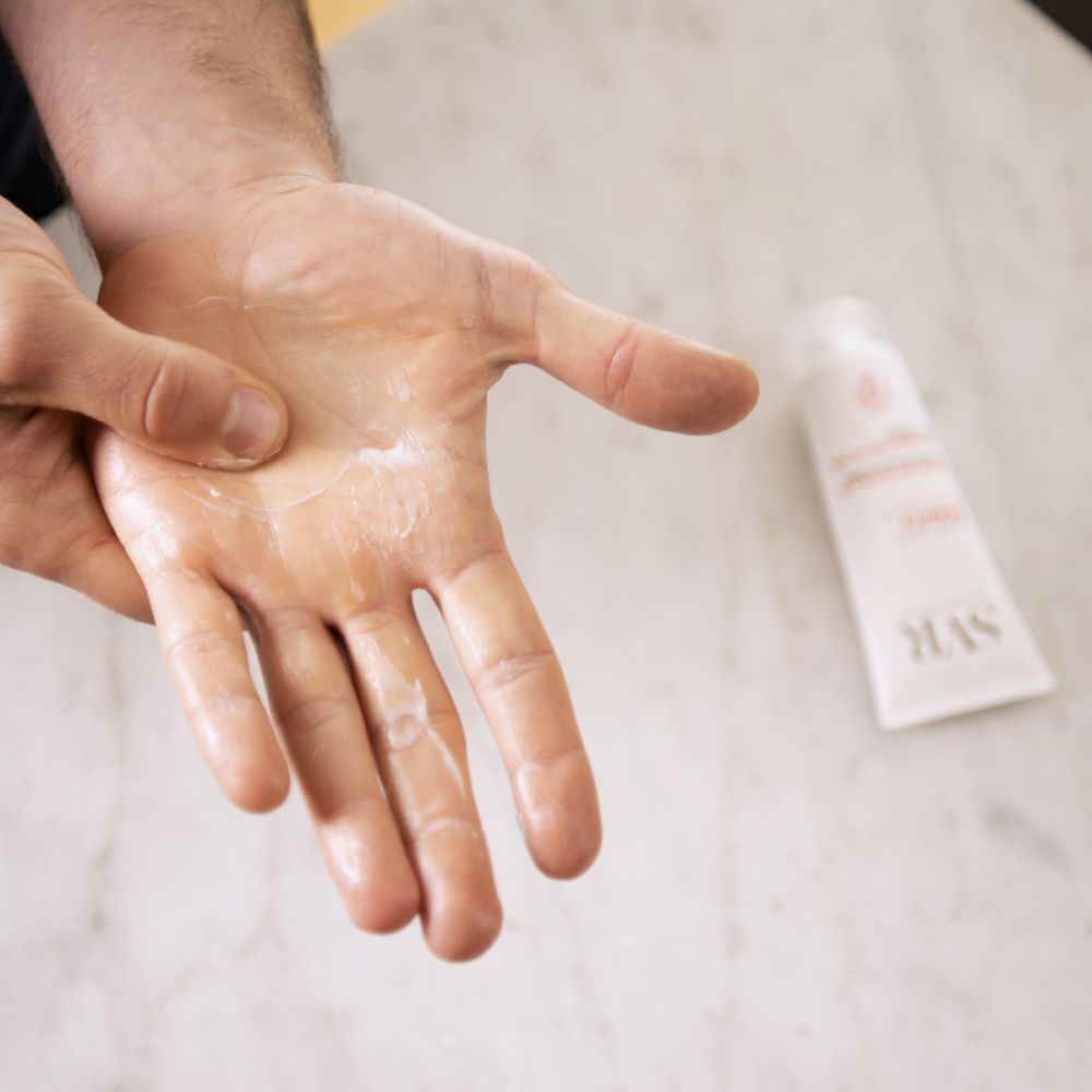 Main avec crème, à côté un tube de SVR Cicavit+ Crème Mains. Crème sur la paume.