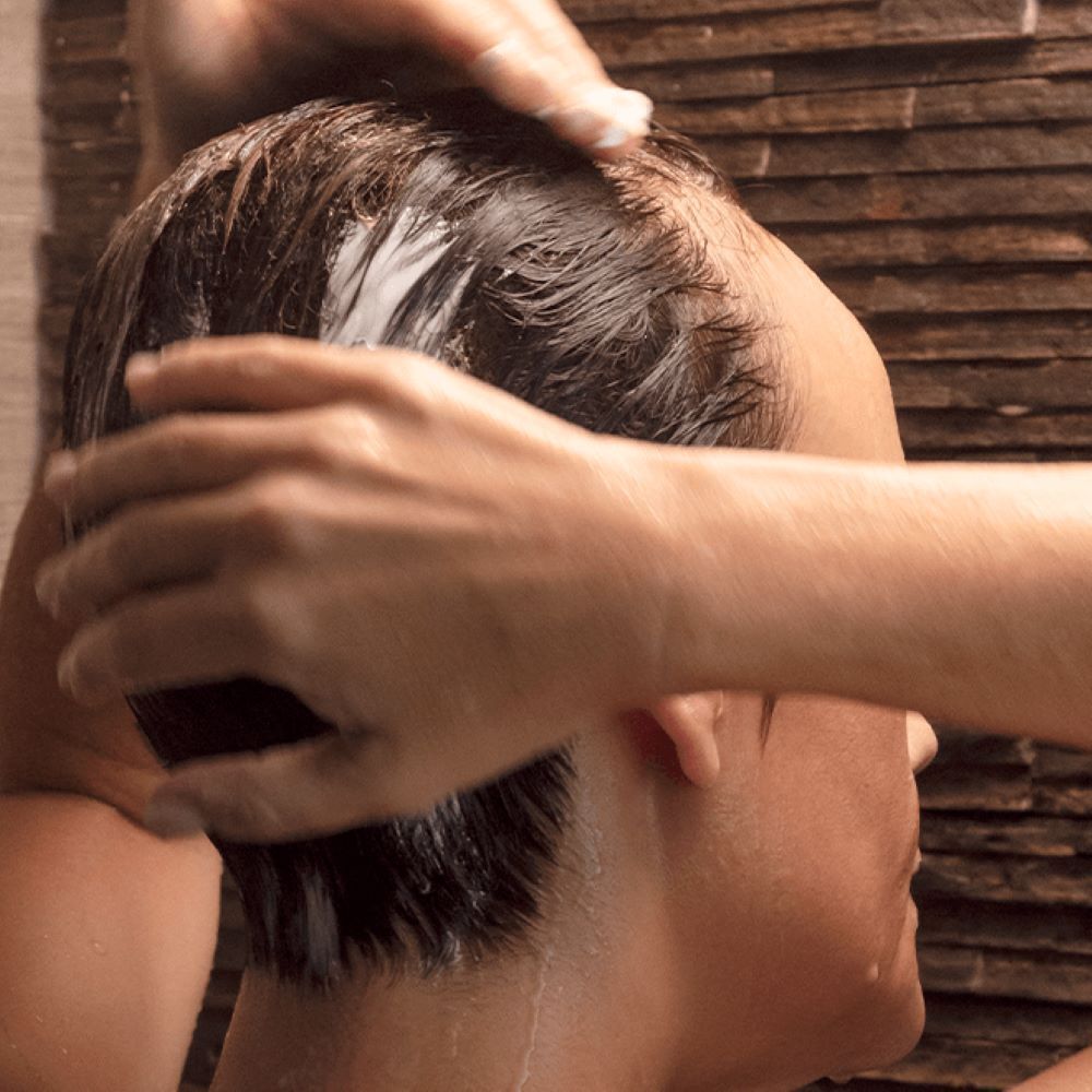 Personne avec cheveux mouillés. Mains massant la tête. Fond: mur de pierre.