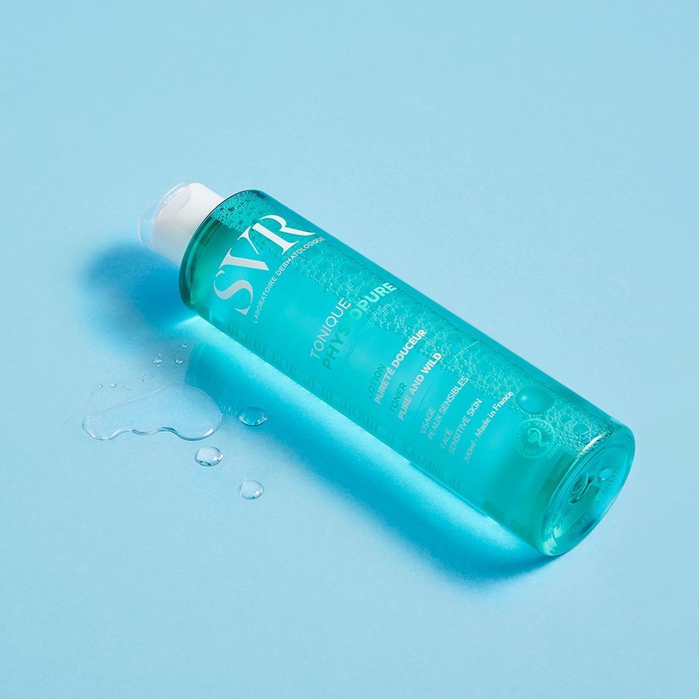 Une bouteille de SVR Physiopure Tonique sur fond bleu. Liquide vert clair dans un flacon cylindrique avec bouchon blanc. Gouttes d'eau.