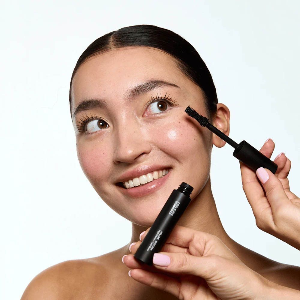 Femme avec applicateur de mascara. Une autre tient le tube de mascara. Nom du produit visible.