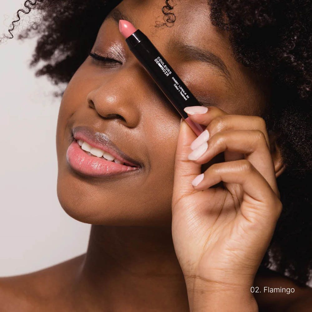 Femme tenant Couleurs de Noir Chubby Lipbalm 02 Flamingo devant l'œil. Rouge à lèvres sur les lèvres. Texte sur le produit.