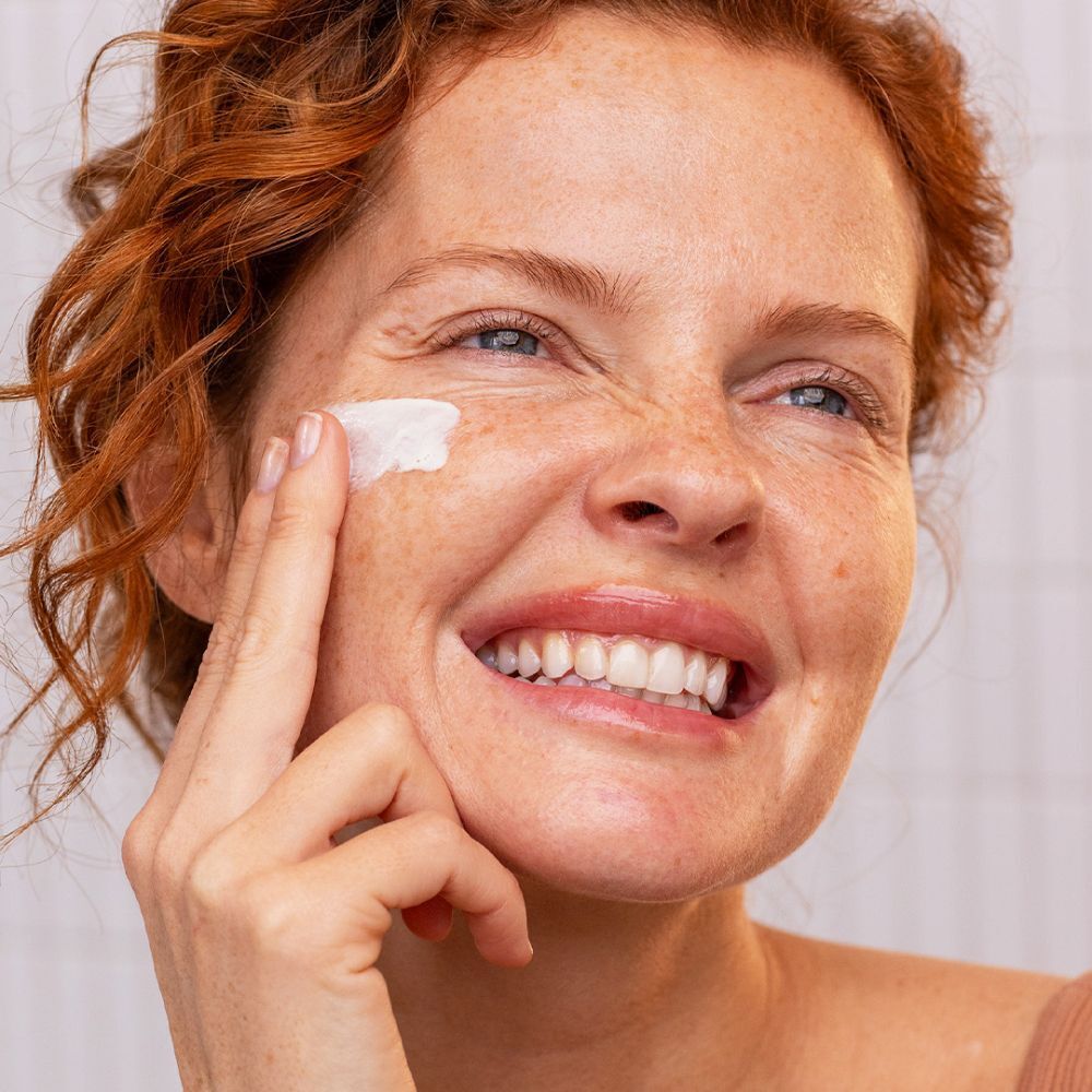 Une femme applique de la crème sur son visage. Elle sourit. Des carreaux blancs sont en arrière-plan.