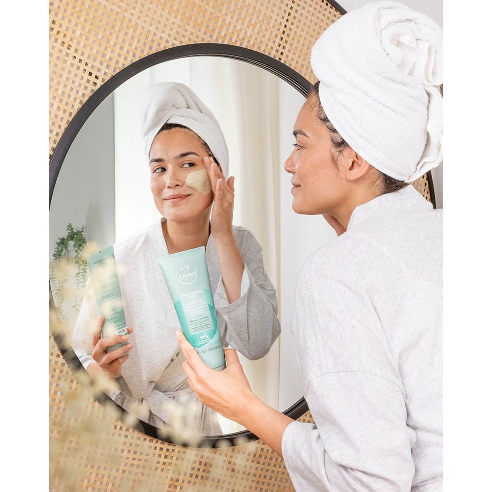 Vrouw brengt Laino Pâte Argile Verte aan op haar gezicht. Ze bekijkt zichzelf in de spiegel. Een product is zichtbaar in haar hand.
