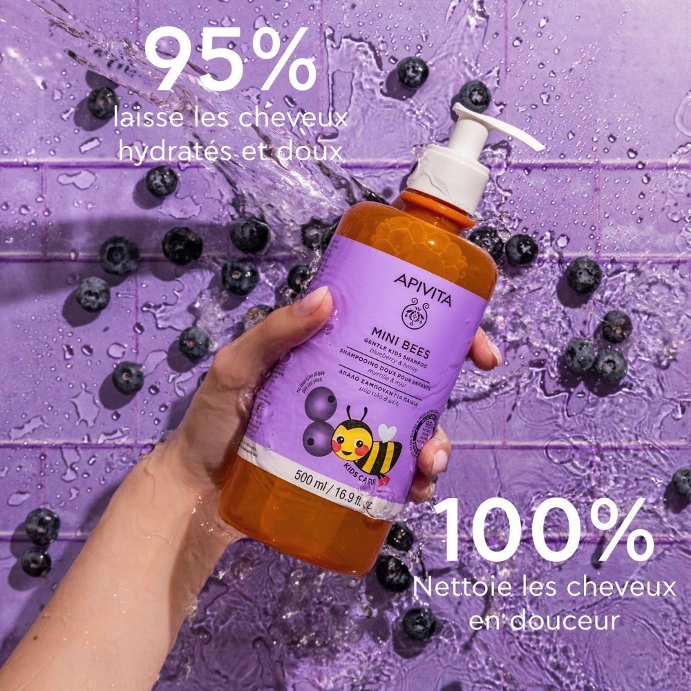Main tenant un flacon de shampooing Apivita Mini Bees. Étiquette violette, nom du produit, abeille. Myrtilles en arrière-plan.