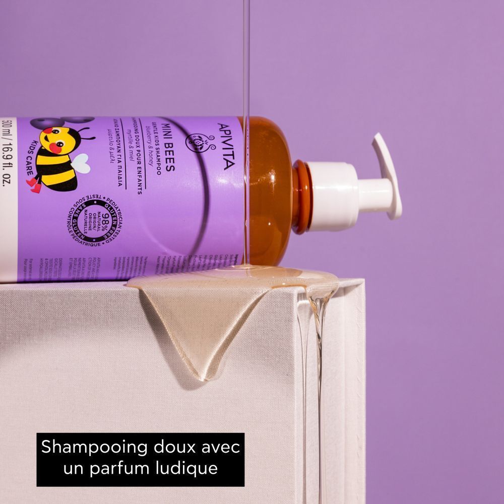 Flacon de shampooing Apivita Mini Bees couché. Étiquette violette, nom du produit, abeille. Liquide qui coule.