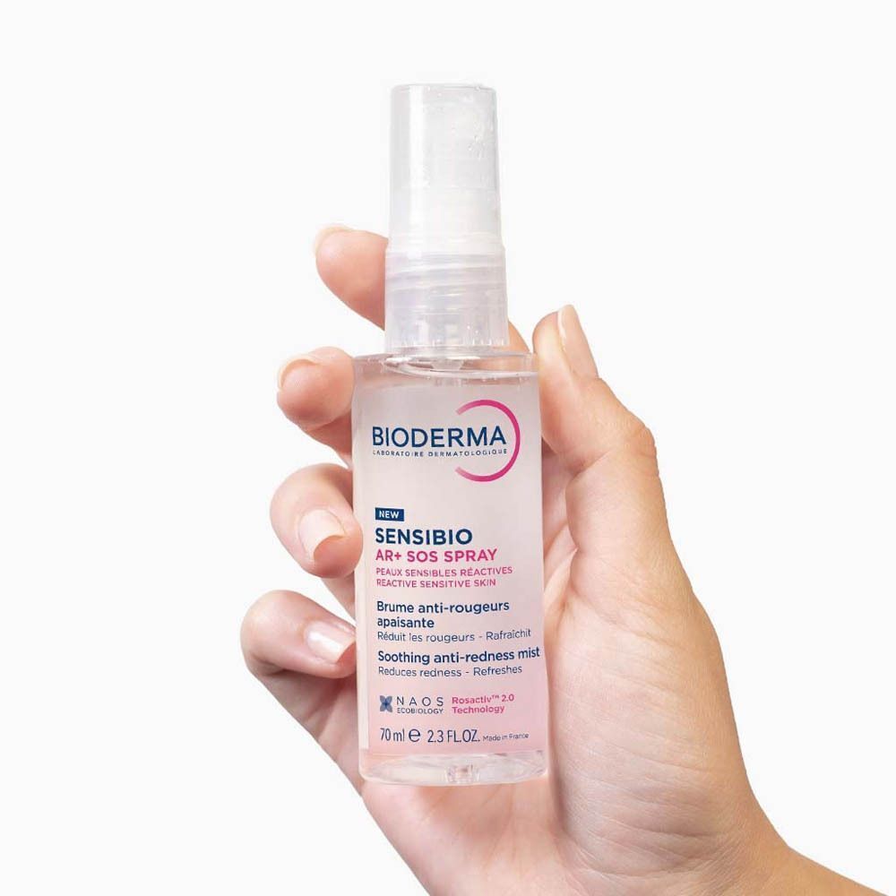 Main tenant Bioderma Sensibio AR+ SOS Spray. Flacon blanc avec vaporisateur, étiquette rose, texte.