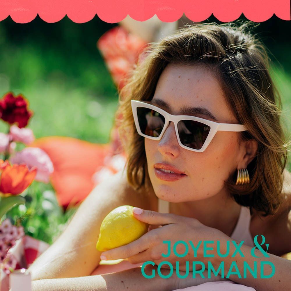 Femme avec des lunettes de soleil allongée dans l'herbe. Texte: 'JOYEUX & GOURMAND'.