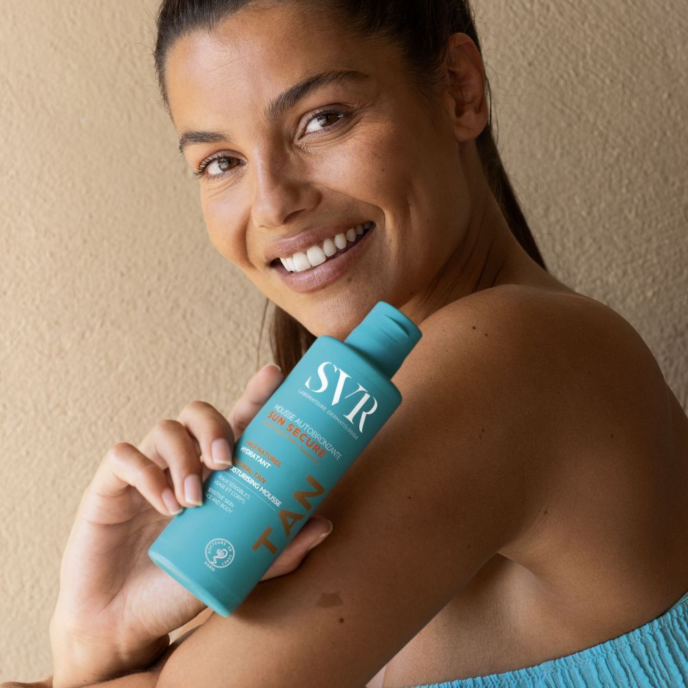Een vrouw die een turquoise flacon SVR Sun Secure Mousse Autobronzante vasthoudt.