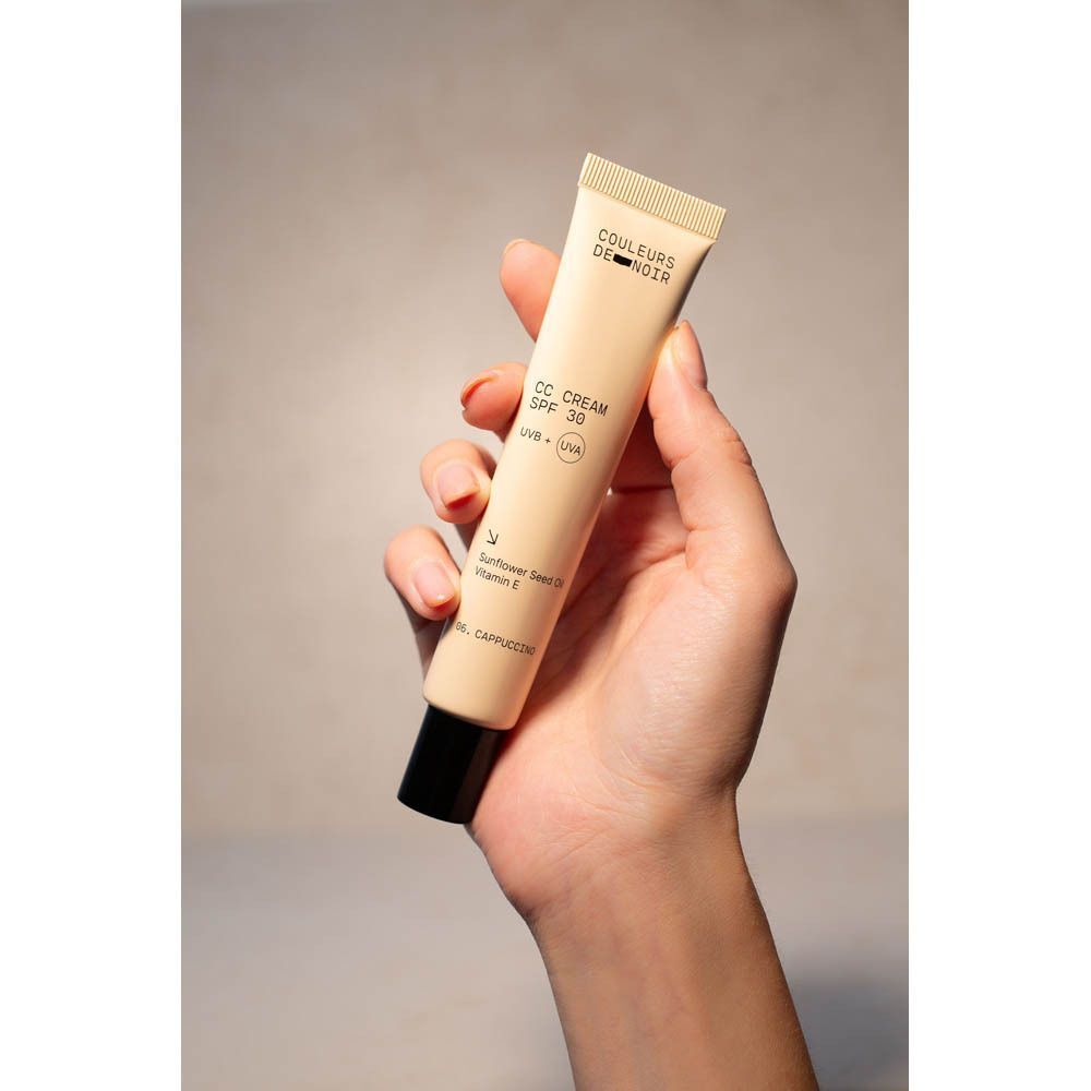 Une main tient un tube de CC Cream. Inscription sur le tube: Couleurs de Noir, CC Cream SPF 30, UVB + UVA, 06 Cappuccino.