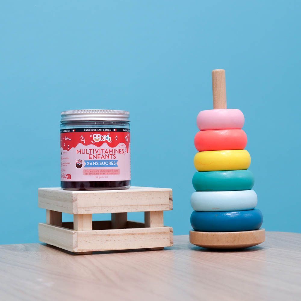 Pot MiumLab Kids Multivitamines Enfants op houten voet. Kleurrijk stapelspeelgoed ernaast. Blauwe achtergrond.