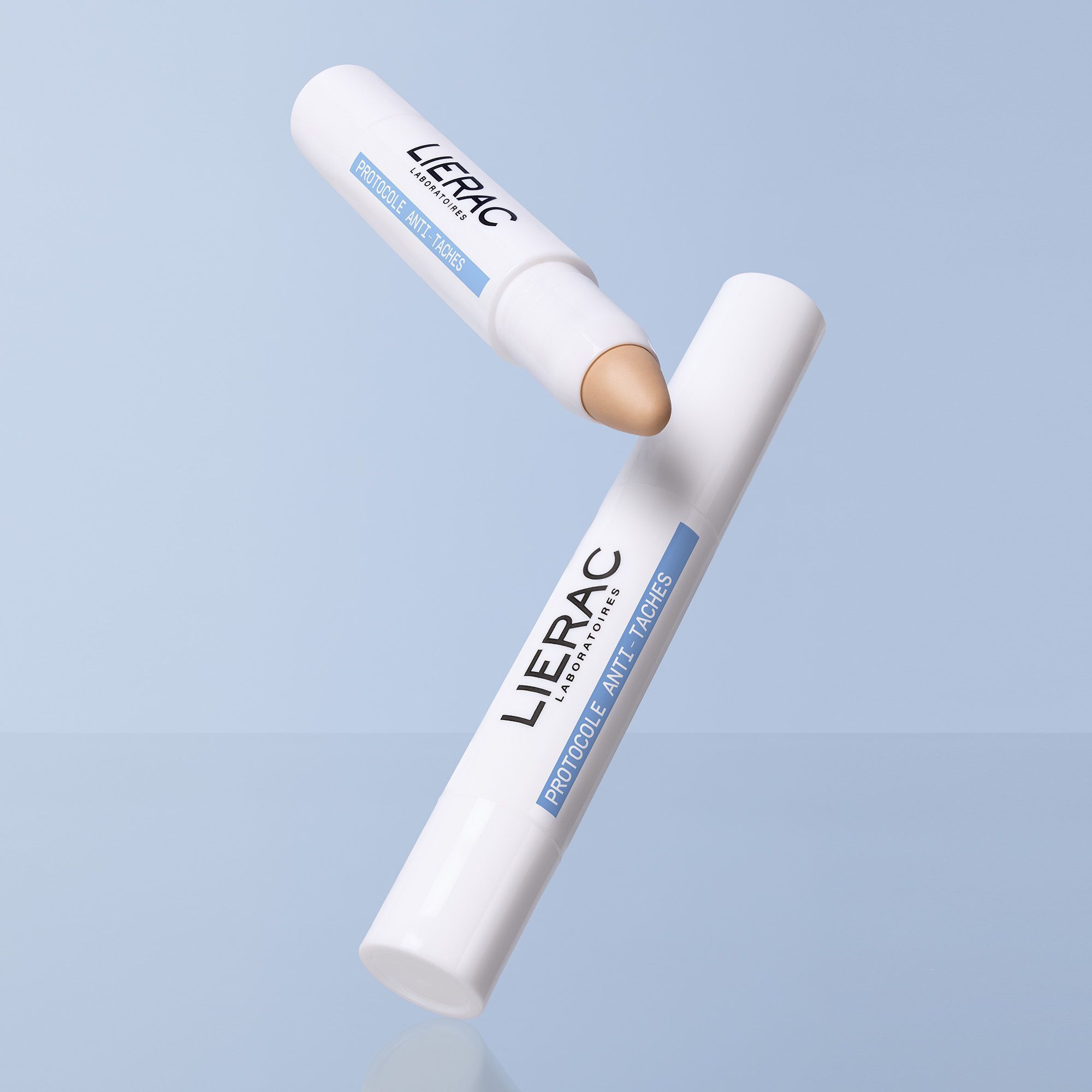 Deux sticks, un avec embout beige, flottent sur fond bleu. Inscription: Lierac, Protocole Anti-Taches.