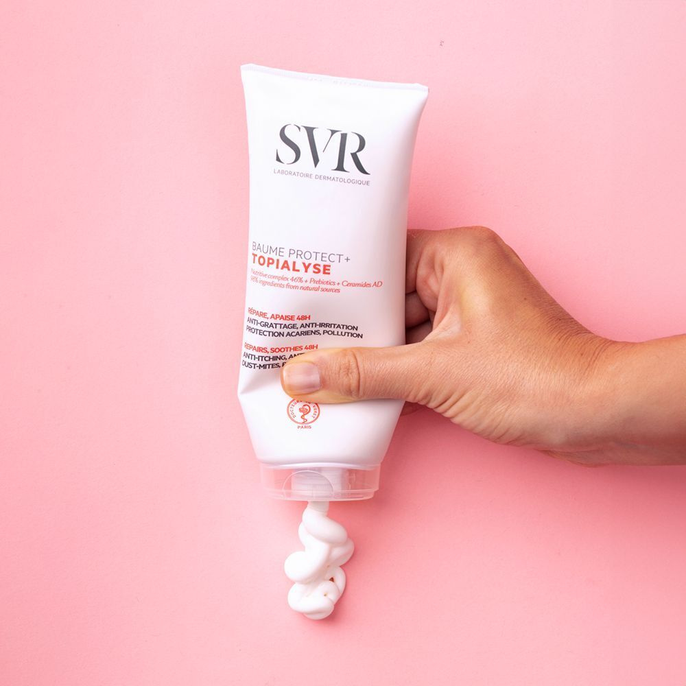 Main tenant un tube de SVR Topialyse Baume Protect+, de la crème sort. Fond rose.