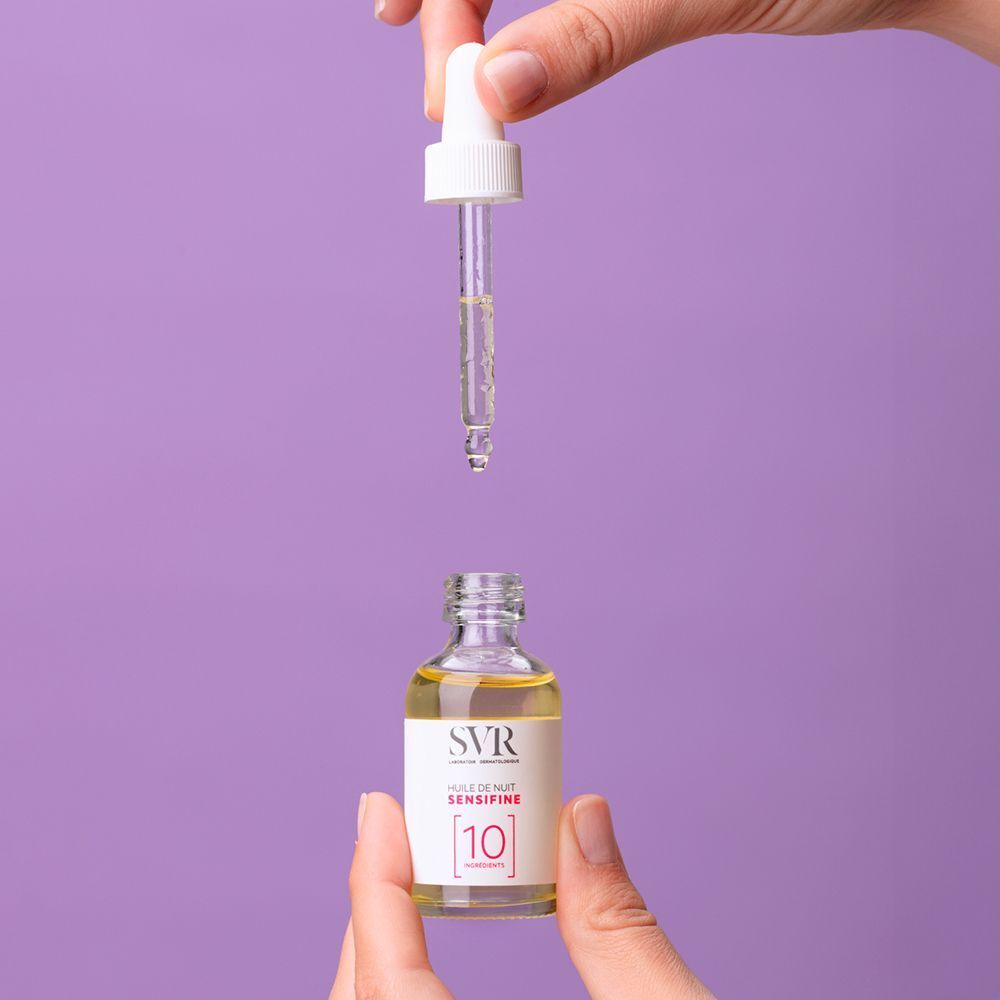 Une main tient un flacon SVR Sensifine [10] Huile de Nuit, l'autre une pipette. La pipette dépose du liquide dans le flacon.