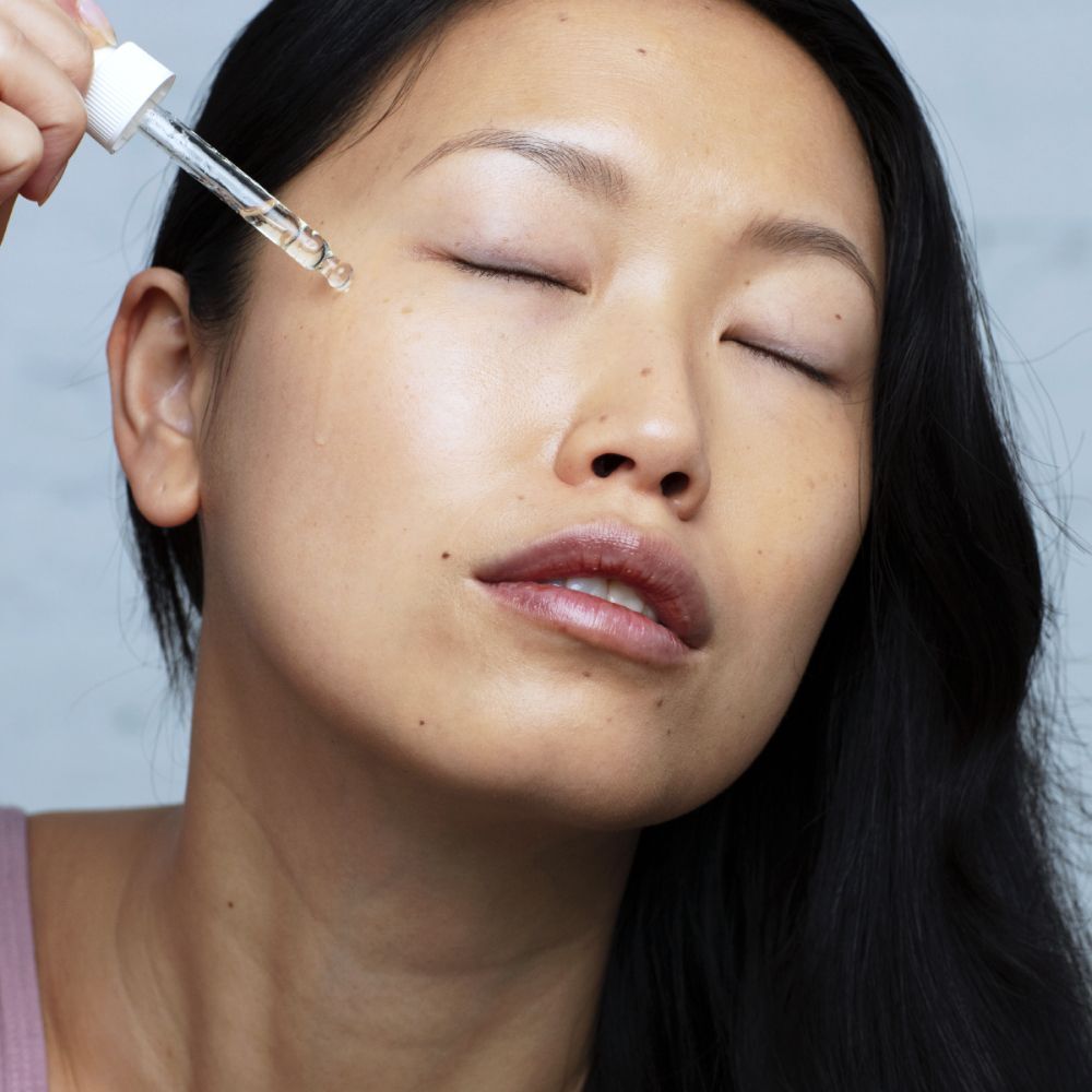 Vrouw met gesloten ogen. Een pipet druppelt vloeistof op de huid. Productlabel niet zichtbaar.