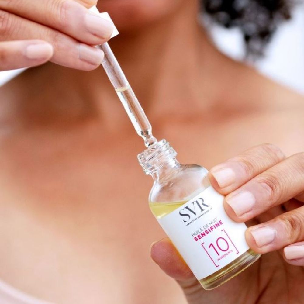 Handen houden een fles en pipet SVR Sensifine [10] Huile de Nuit vast. Pipet in fles. Productlabel zichtbaar.