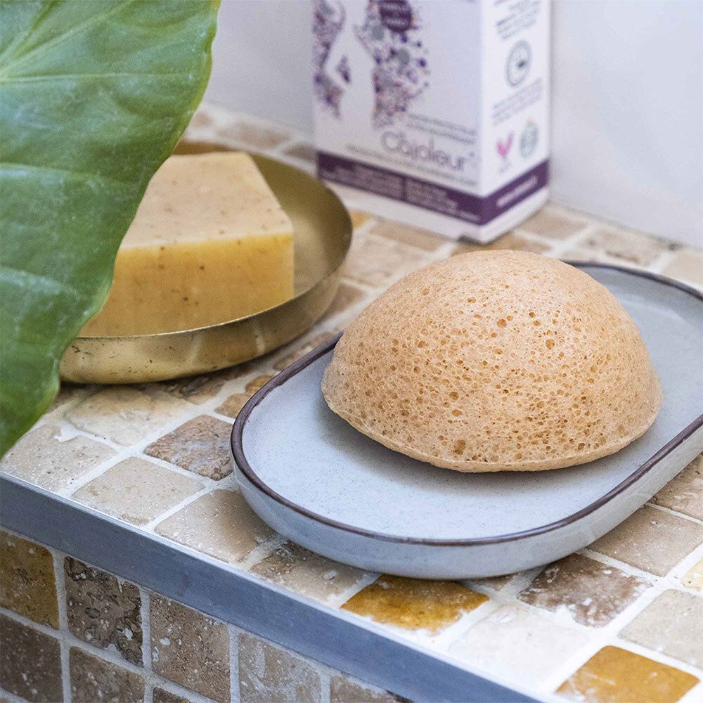 Une éponge konjac ronde et orange sur un porte-savon à côté d'un morceau de savon.
