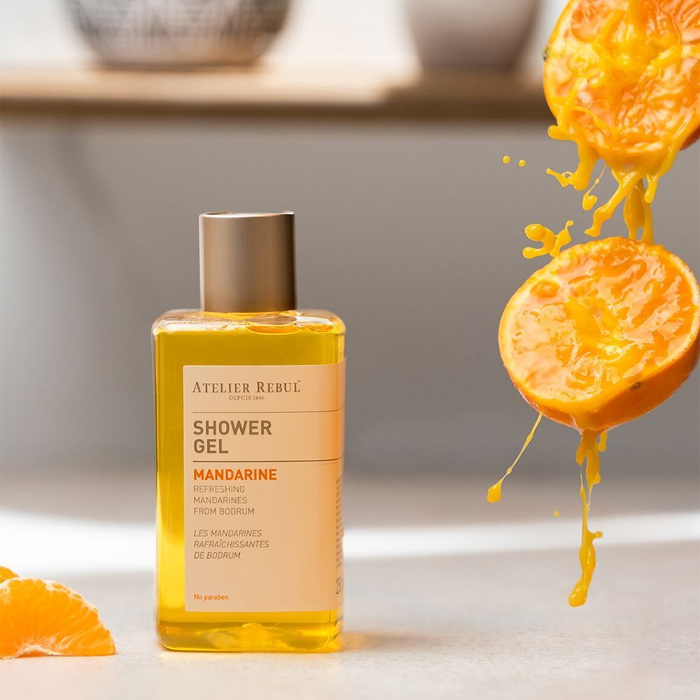 Een fles Atelier Rebul douchegel "Mandarine" naast gesneden mandarijnen en sapspetters.
