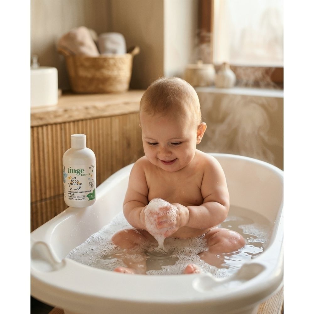 Un bébé souriant assis dans une baignoire pleine de mousse, à côté d'un flacon d'huile de bain Tinge.