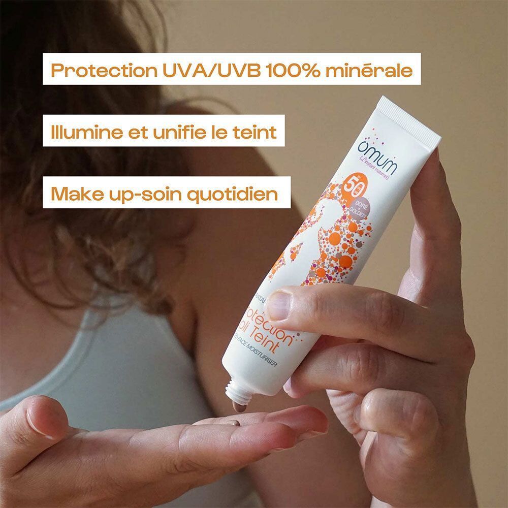 Une main tient un tube de omum Protection Joli Teint SPF 50, appliqué sur l'autre main.