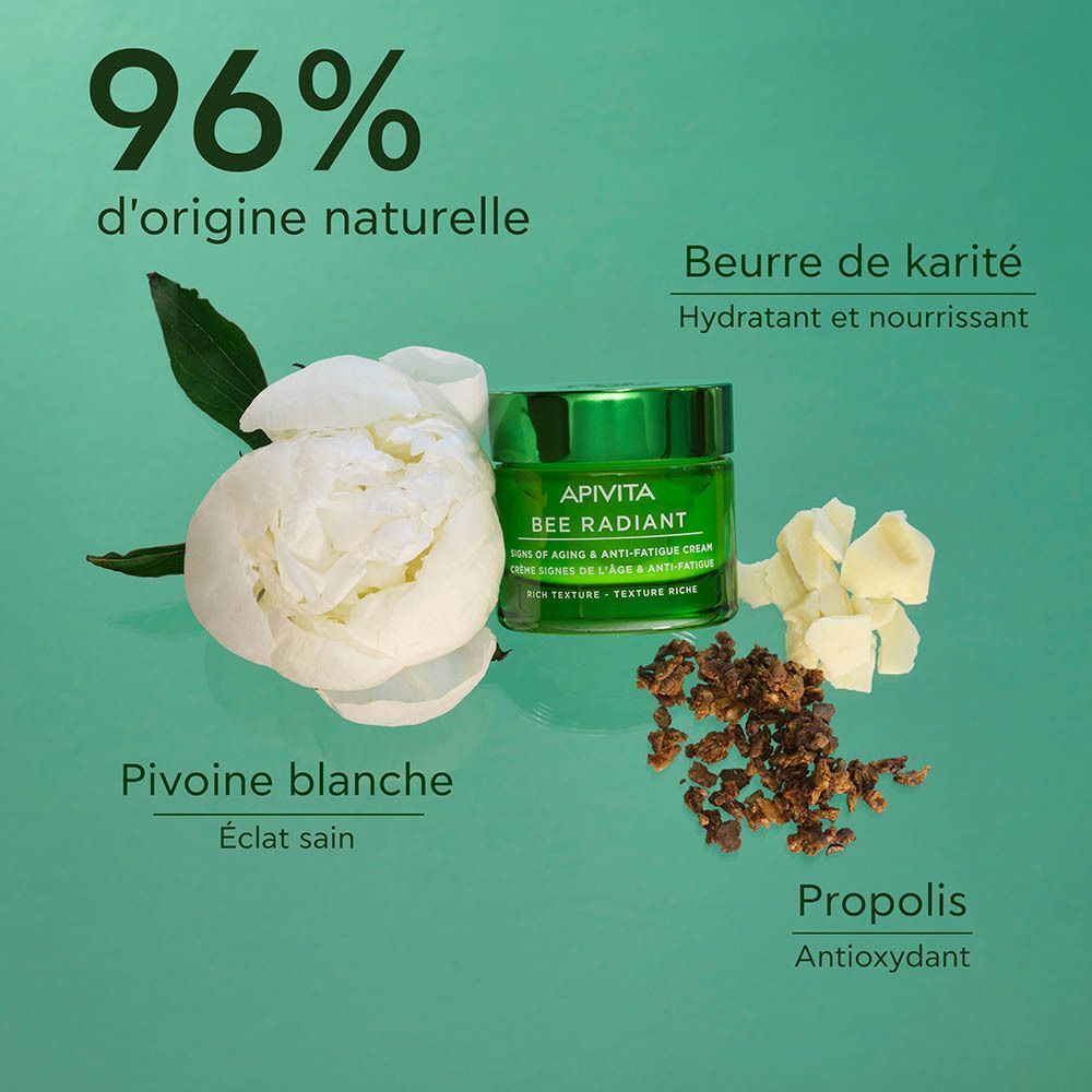 Un pot vert de crème Apivita Bee Radiant entouré de fleurs blanches, de beurre de karité et de propolis.