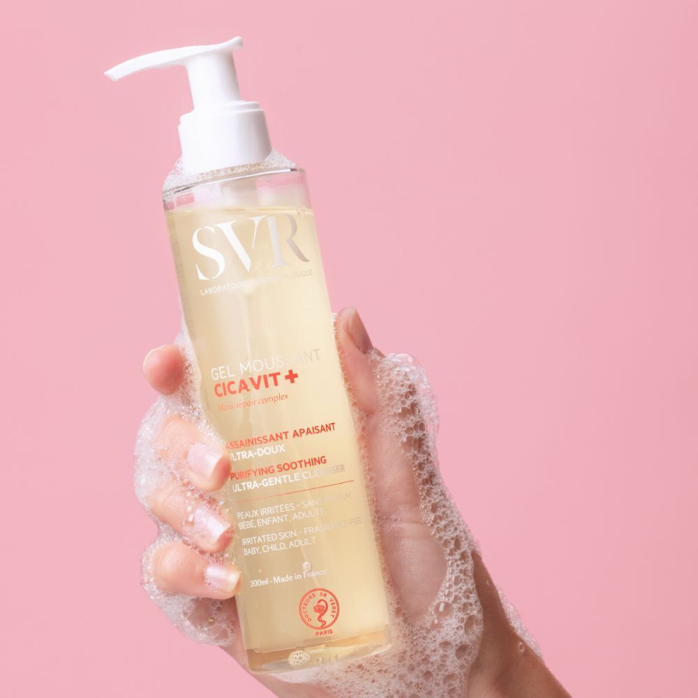 Een hand houdt een fles SVR Cicavit+ Gel Moussant vast, die schuim produceert.