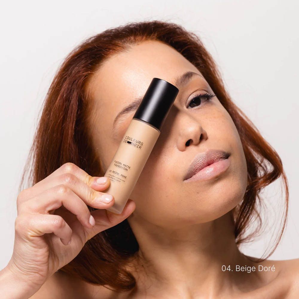 Une femme tient un flacon de fond de teint COULEURS DE NOIR Fusion Glow, teinte 04 Beige Doré.