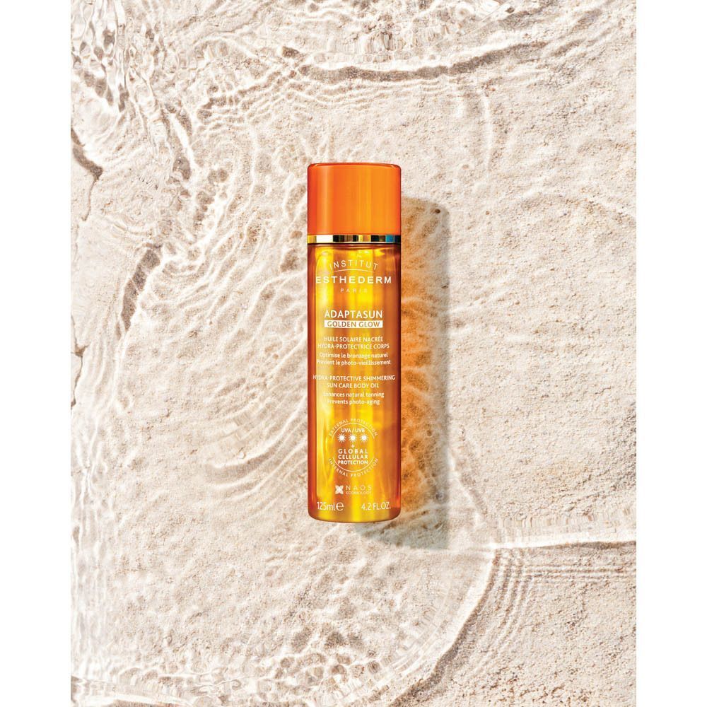 Huile Solaire Nacrée Hydra-Protectrice Corps Institut Esthederm Golden Glow sur fond de sable avec des ondulations d'eau.
