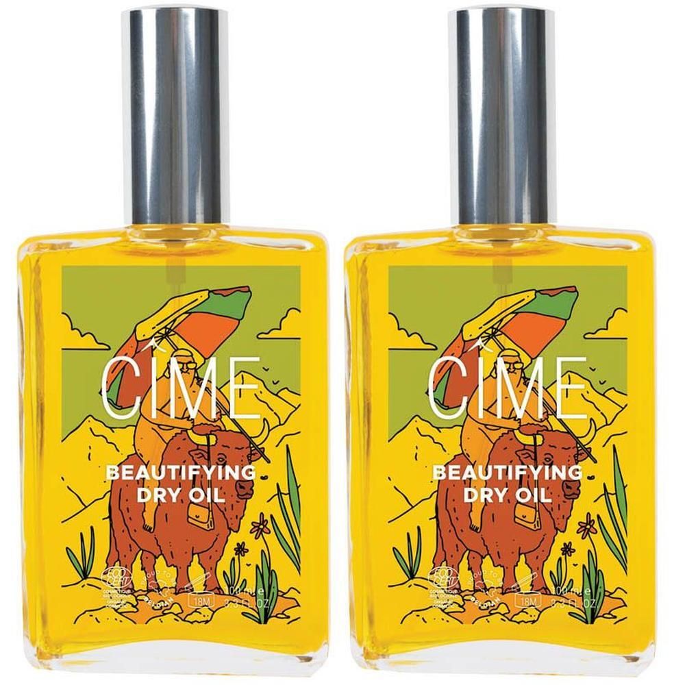 Twee flessen Cîme Huile sèche sublimante met zilveren doppen. Gele etiketten tonen een woestijnlandschap met een figuur op een buffel.