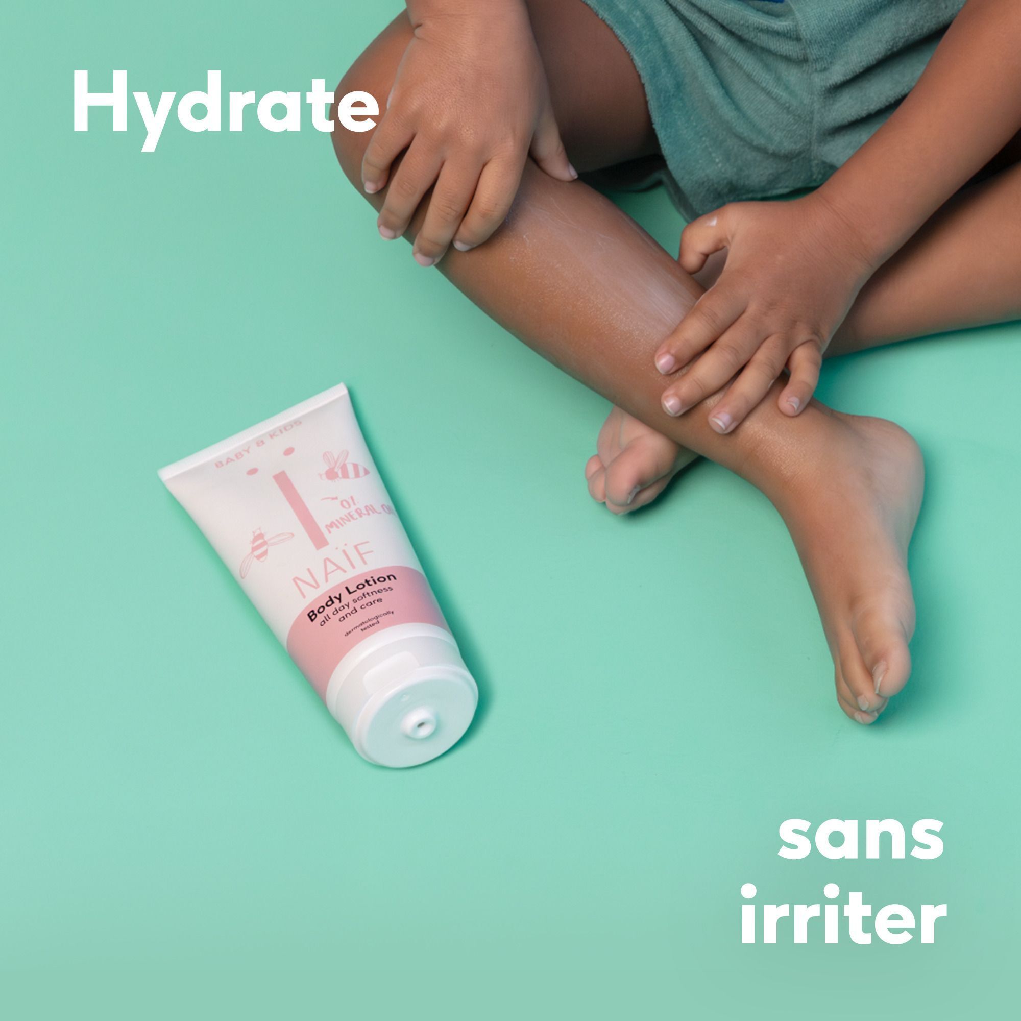 Un enfant assis, un tube de lotion corporelle Naïf à côté de sa jambe.