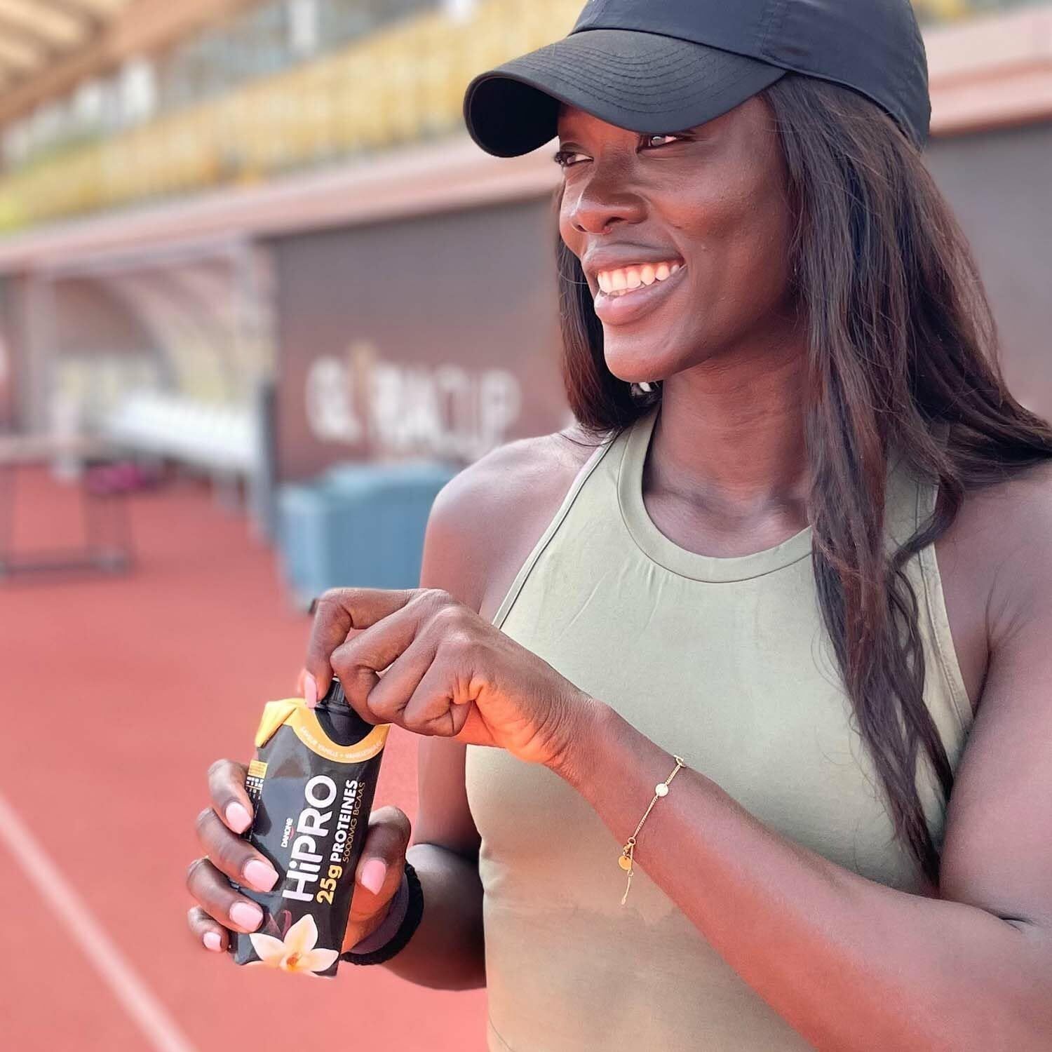 Een vrouw opent een flesje Danone HiPRO Drink Vanille op een sportveld.