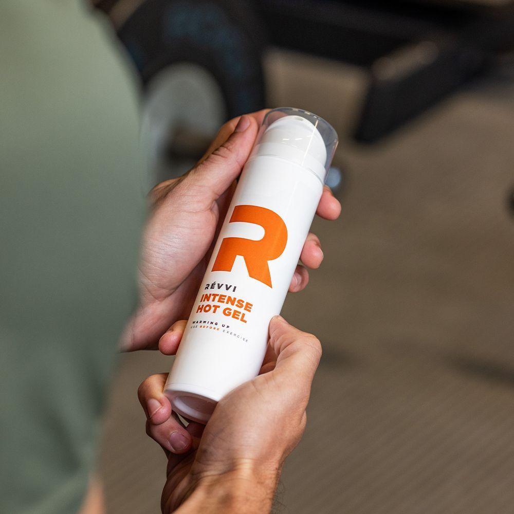 Handen houden een wit pompflesje vast met oranje 'R'-logo en opschrift 'RÉVVI INTENSE HOT GEL'.