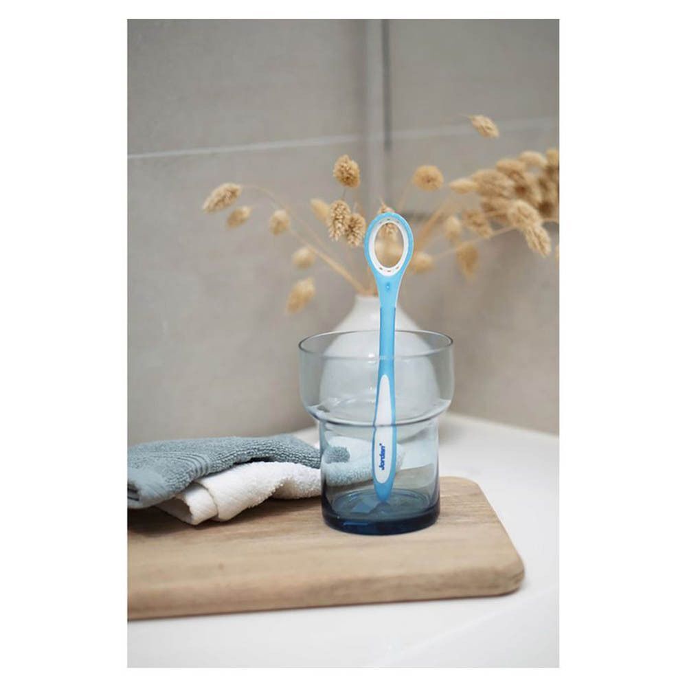 Een Jordan Clinic tongreiniger in een glas op een houten schaal, naast handdoeken.