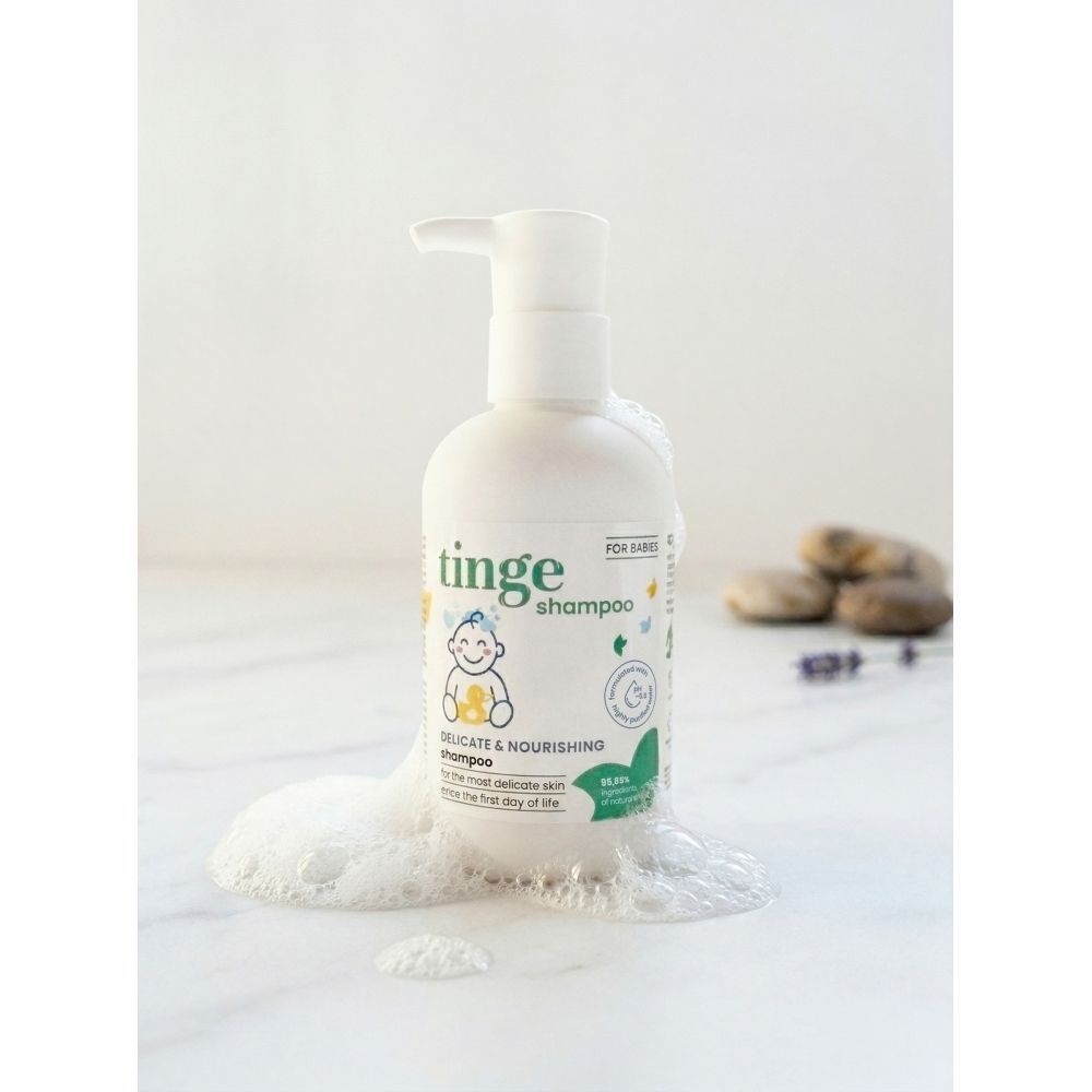 Un flacon blanc de shampooing pour bébé Tinge avec distributeur à pompe, à côté d'un peigne et d'une brosse en bois sur un tissu vert.