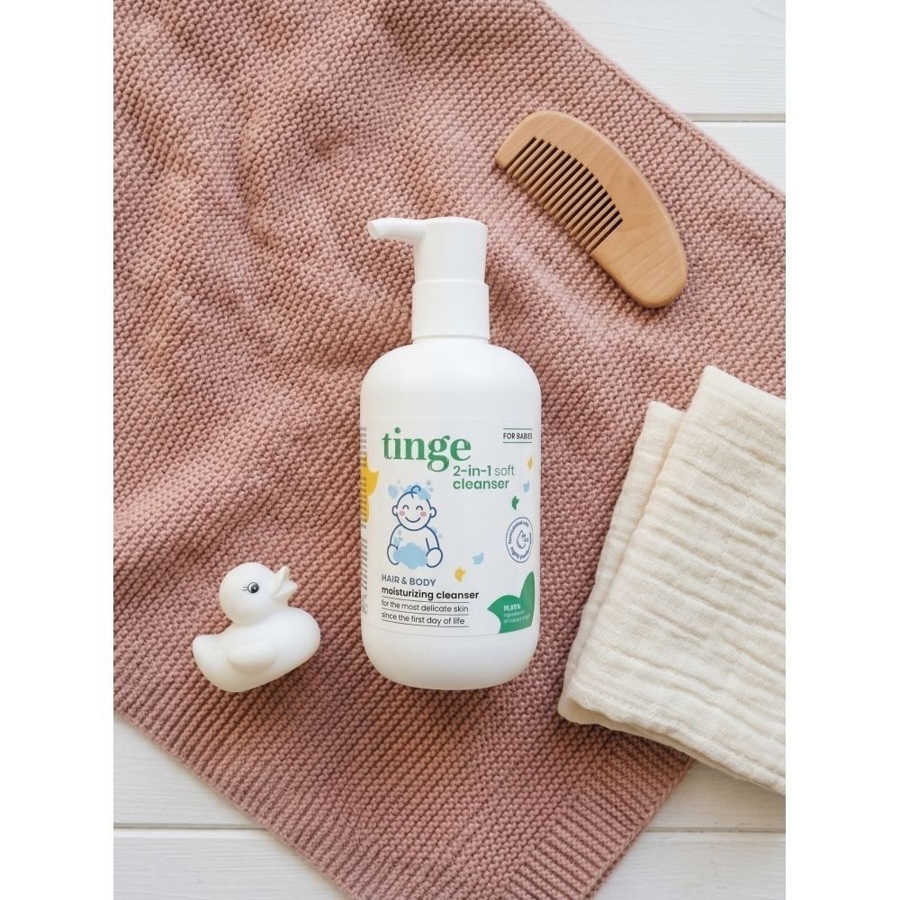 Tinge Gel Lavant 2-en-1 pour bébés sur une serviette rose avec canard en caoutchouc et peigne en bois.
