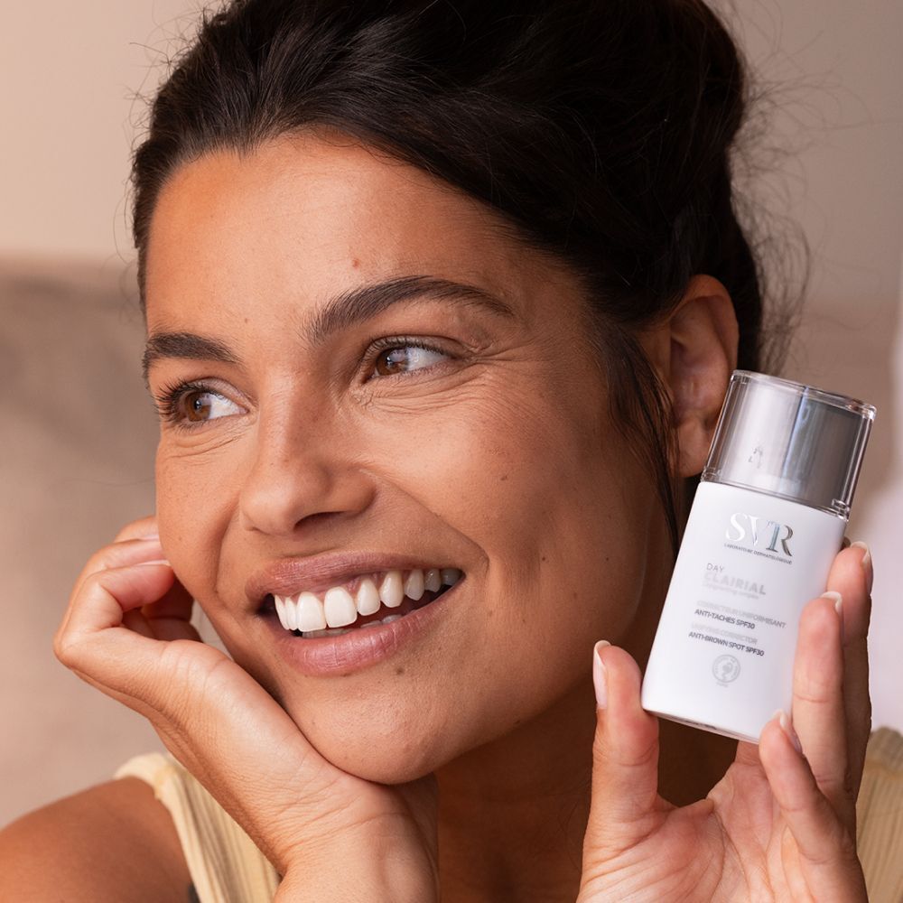 Vrouw die een witte flacon SVR Clairial Day SPF30 met pompdos vasthoudt.