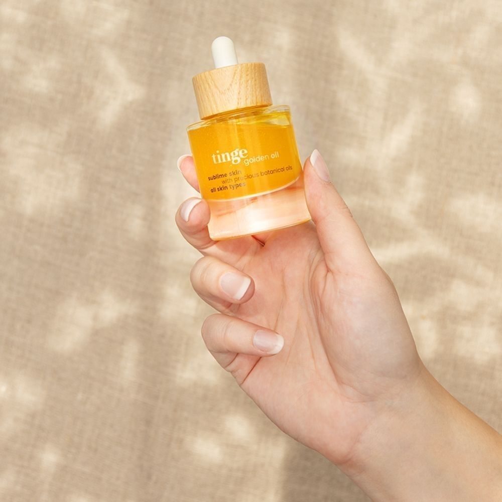 Een hand houdt een fles Tinge Golden Oil vast met pipet en houten dop.