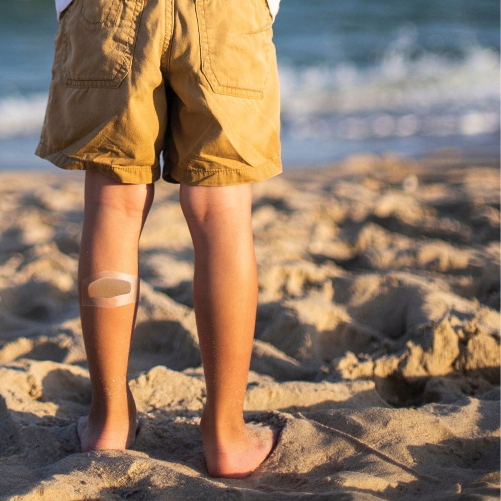 Un enfant sur la plage avec un pansement Nexcare Aqua Clear Maxi sur le tibia.