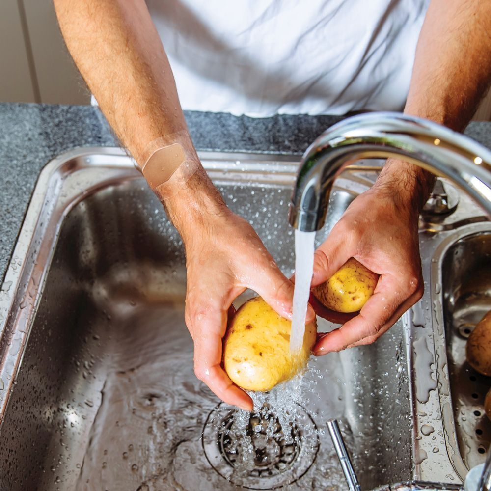 Homme lavant des pommes de terre sous l'eau courante avec un pansement Nexcare Aqua Clear Maxi au poignet.