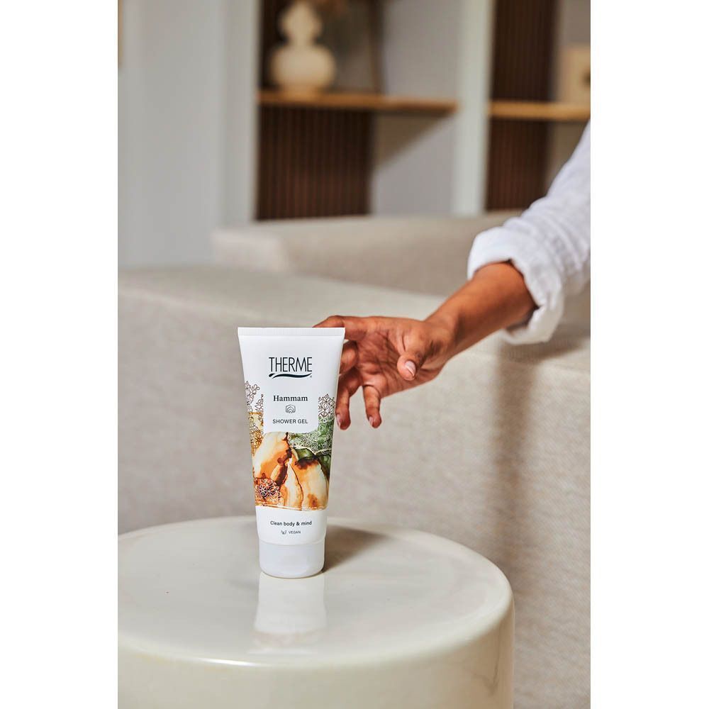 Een hand pakt een tube Hammam douchegel van Therme op een bijzettafel.