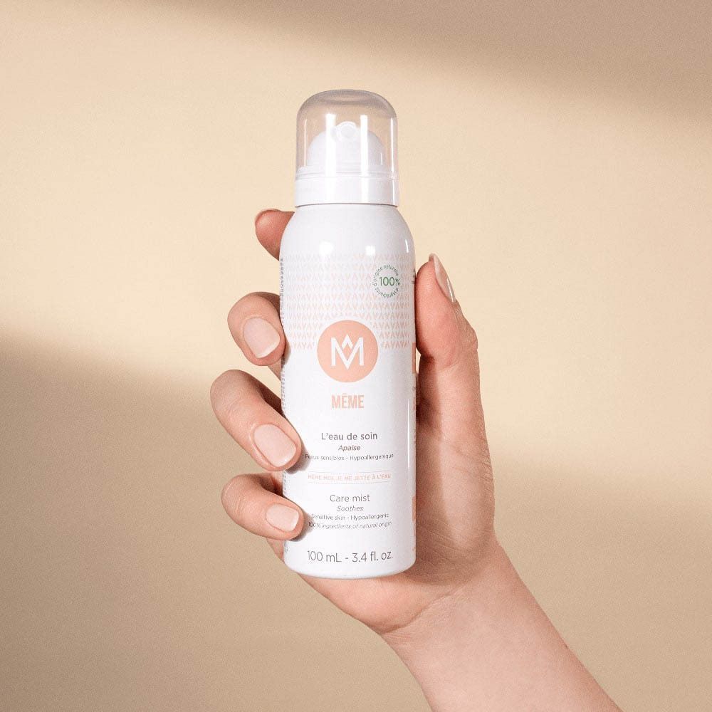 Une main tient un flacon spray MÊME L'eau de soin sur fond neutre.