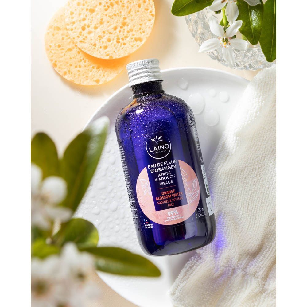 Un flacon bleu Laino Eau de Fleur d'Oranger sur une surface blanche avec éponges et serviette.