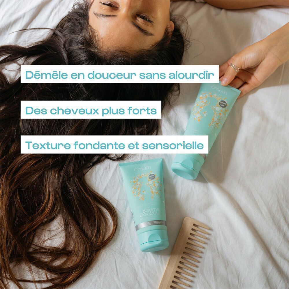 Deux tubes Omum Mon Baume Douceur Après-Shampoing près de longs cheveux sombres et d'un peigne en bois.