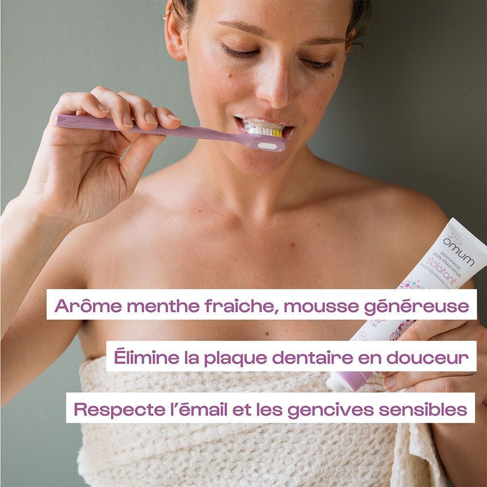 Une femme se brosse les dents avec une brosse à dents, tenant un tube de dentifrice Omum.