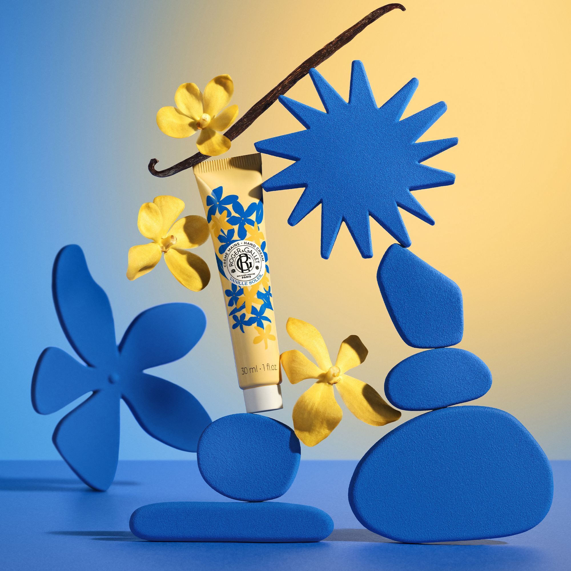 Een tube handcrème met Vanille Soleil-motief, omringd door blauwe vormen en gele bloemen.