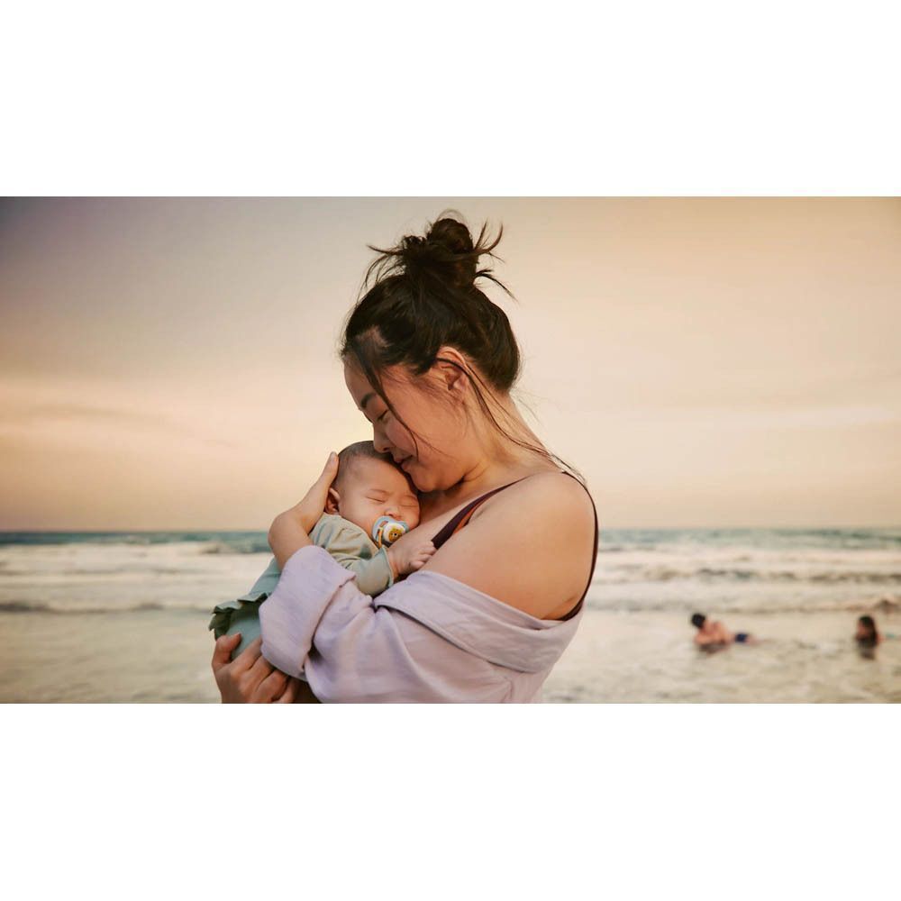 Moeder houdt baby vast op het strand. Baby heeft een fopspeen met leeuwenmotief in de mond.