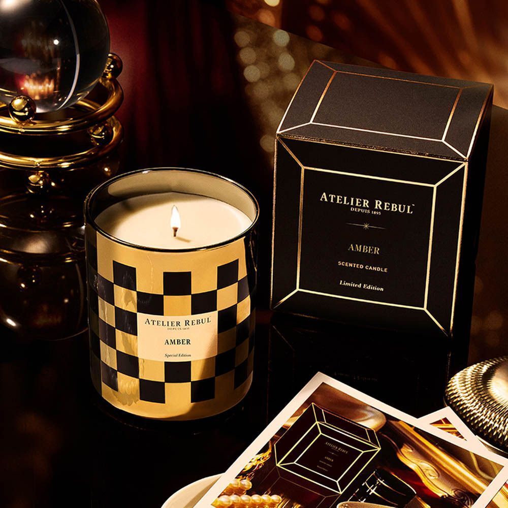 Bougie parfumée Atelier Rebul Ambre allumée, motif damier or et noir, à côté de sa boîte.